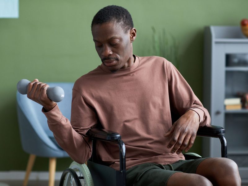 Waist up portrait of African American man with disability in wheelchair exercising at home using dumbbell.
