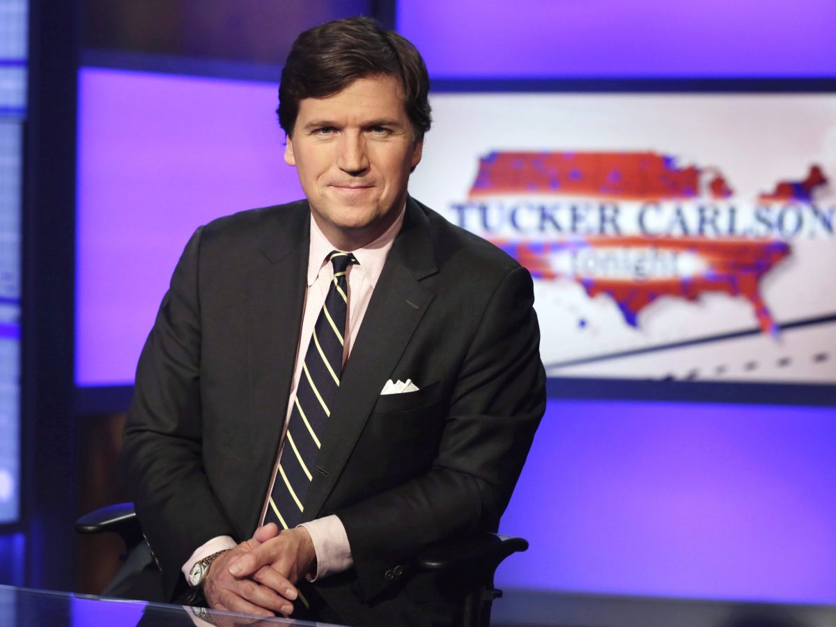 Tucker Carlson poses in suit and tie in a Fox News Channel studio with his shows logo on screen in background.